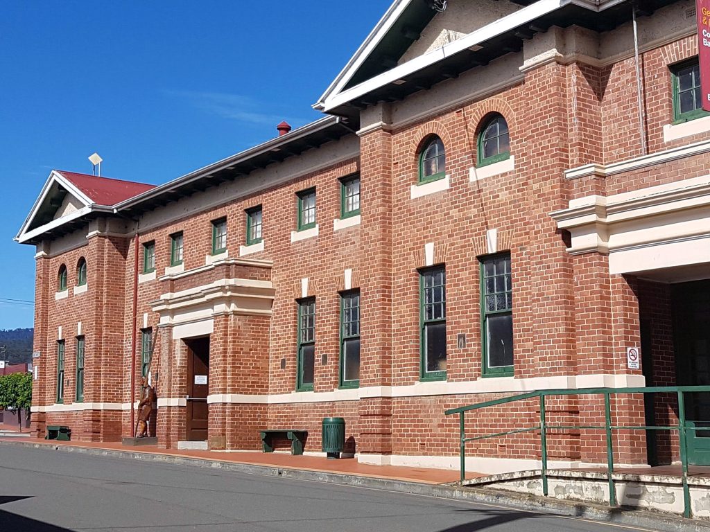 A two-story red brick building with green-framed windows and a red roof under a clear blue sky. The facade shows arched windows and a prominent entrance with white detailing. A green metal railing and sidewalk are in the foreground.