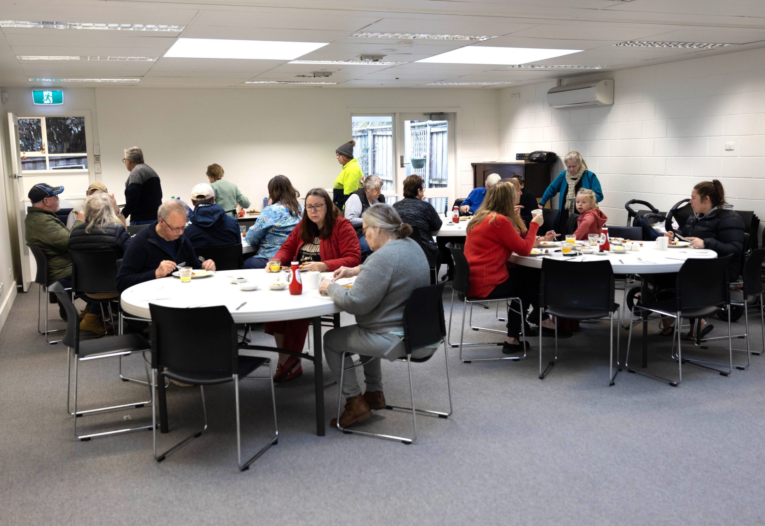 A group of people seated around tables in a community hall, eating and conversing. The room is well-lit with white walls, and there are several round tables with chairs. Some people are standing near the entrance.