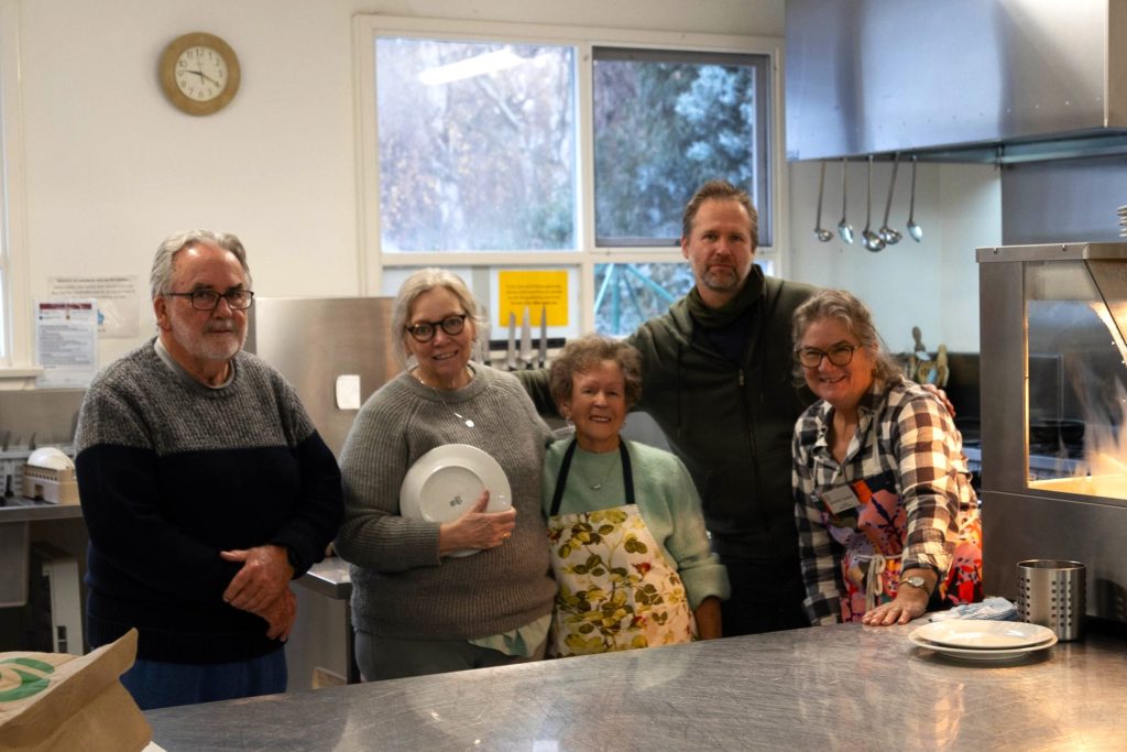A group of five people standing together in a kitchen. They are smiling, with some holding dishes. The kitchen has a window, stainless steel surfaces, and various kitchen equipment. Natural light streams in through the window.
