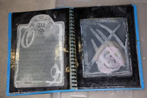 An open childrens scrapbook with a Me page on the left, containing handwritten text and face illustrations. The right page shows a childs colorful drawing of a face with a dark background. The book appears aged and worn.
