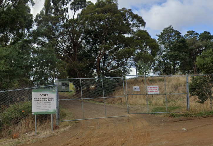 A dirt road leads to a gated entrance surrounded by a chain-link fence. A sign on the left reads Dover Waste Transfer Station and Recycling Centre. Trees and grass line the area, with a partly cloudy sky overhead.