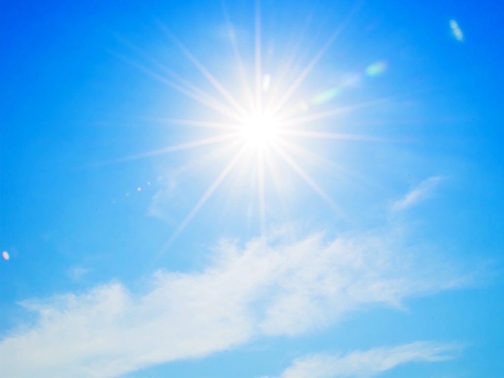 Bright sun with rays shining in a clear blue sky, small wispy clouds scattered below.