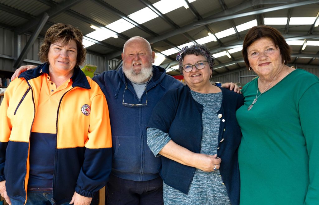Four people standing close together inside a well-lit building. The person on the left is wearing an orange jacket, the man next to them a blue hoodie, the third person a dark cardigan, and the fourth a green top. They are all smiling at the camera.