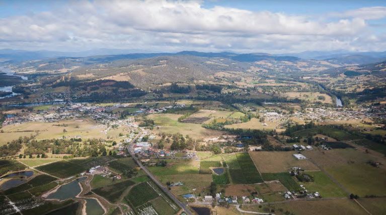 Aerial view of a rural landscape with fields, trees, and a small town. Rolling hills and mountains are visible in the background under a partly cloudy sky. The scene shows a mix of agricultural land and small clusters of buildings.
