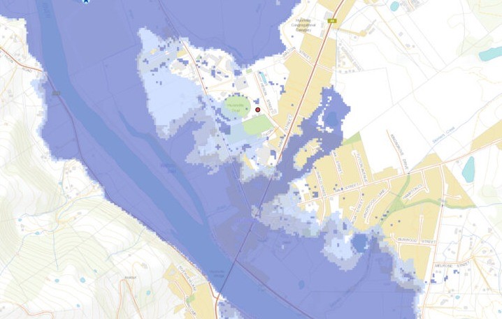 Map depicting a river near a town, with surrounding areas shaded in blue to indicate potential flood zones. Various roads and civic features are highlighted, such as buildings and green spaces.