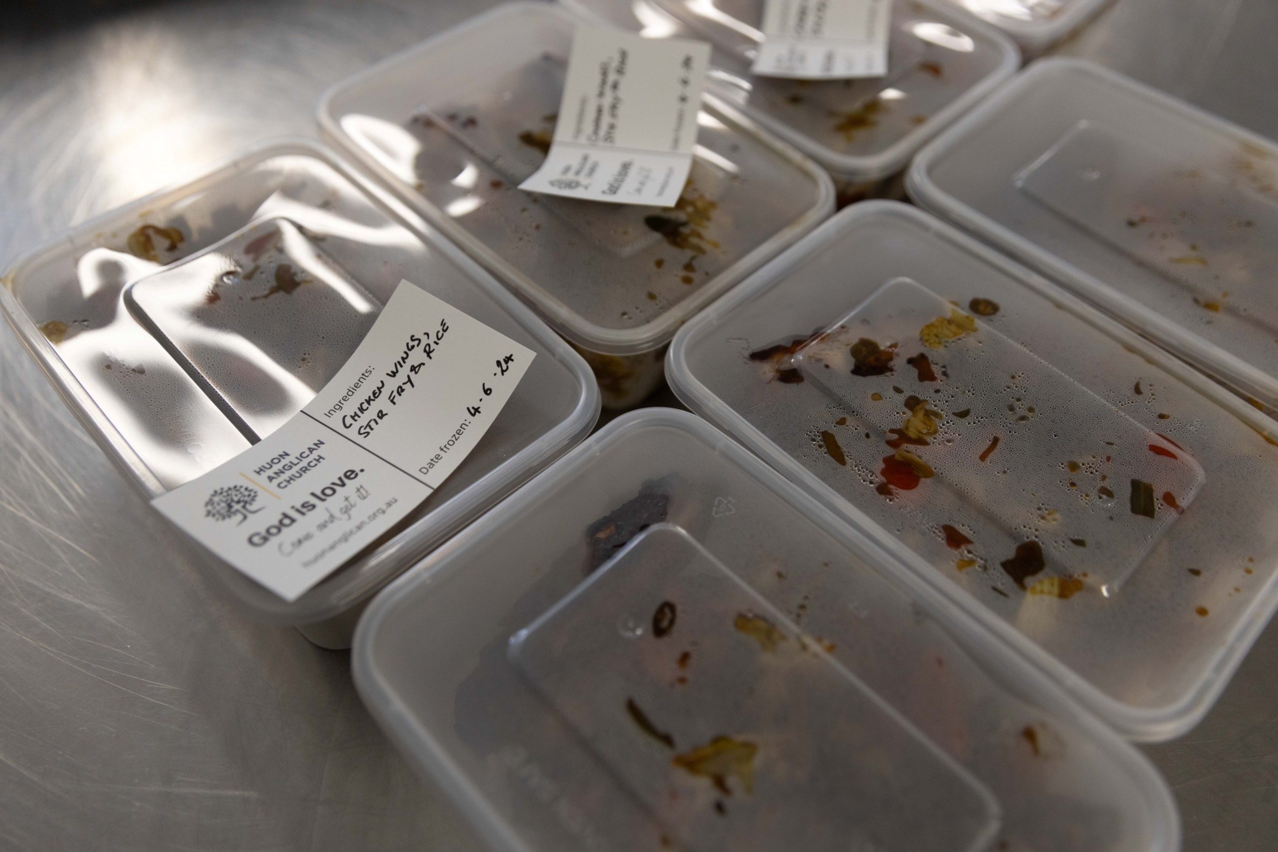 Clear food containers with remnants of meals and labeled with stickers are arranged on a metallic surface. The labels include details like date, meal names, and an organizations logo with the words Gods Love.