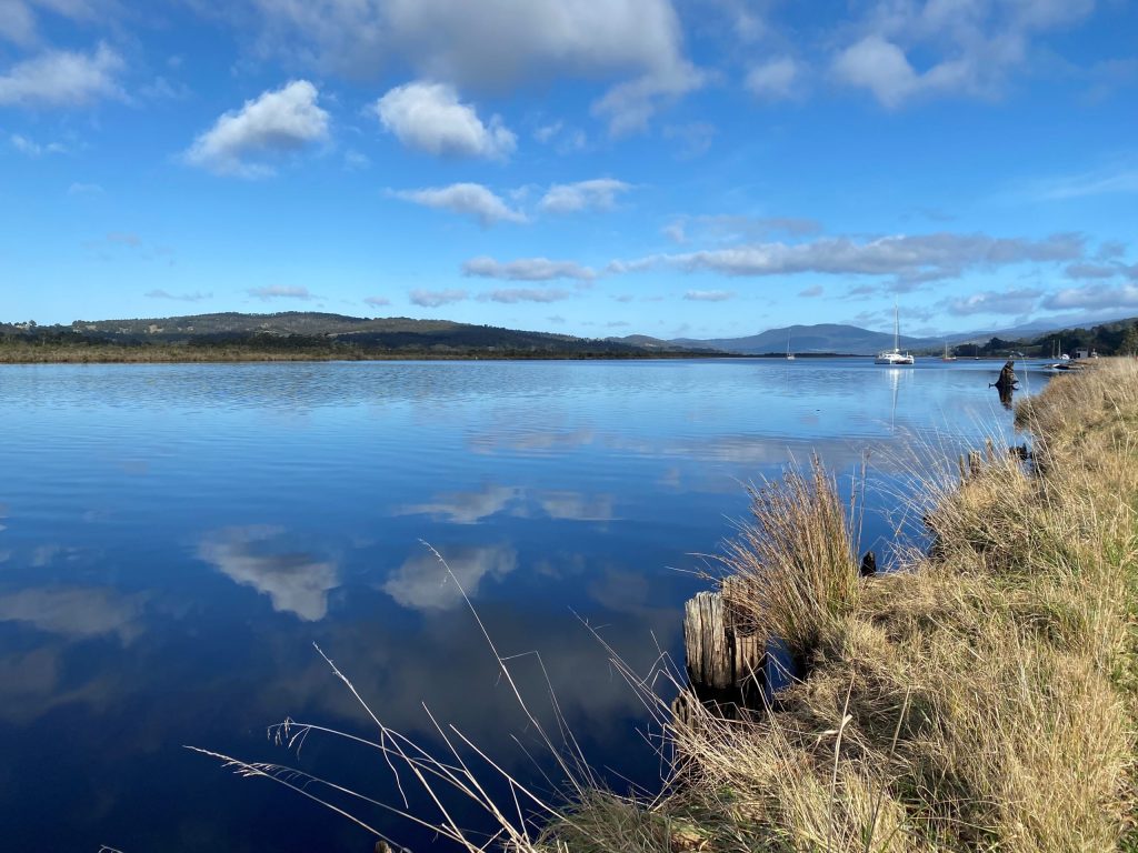 A tranquil lake with a clear blue sky reflected on its surface. Grassy banks line the waters edge, and a sailboat is anchored in the distance. Fluffy clouds are scattered across the sky, and distant hills are visible on the horizon.