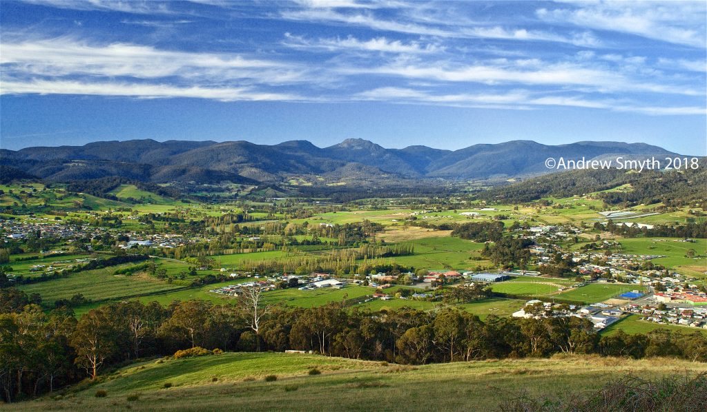 A scenic view of a sprawling green valley with scattered houses, surrounded by forested hills and mountains under a blue sky with streaky clouds.