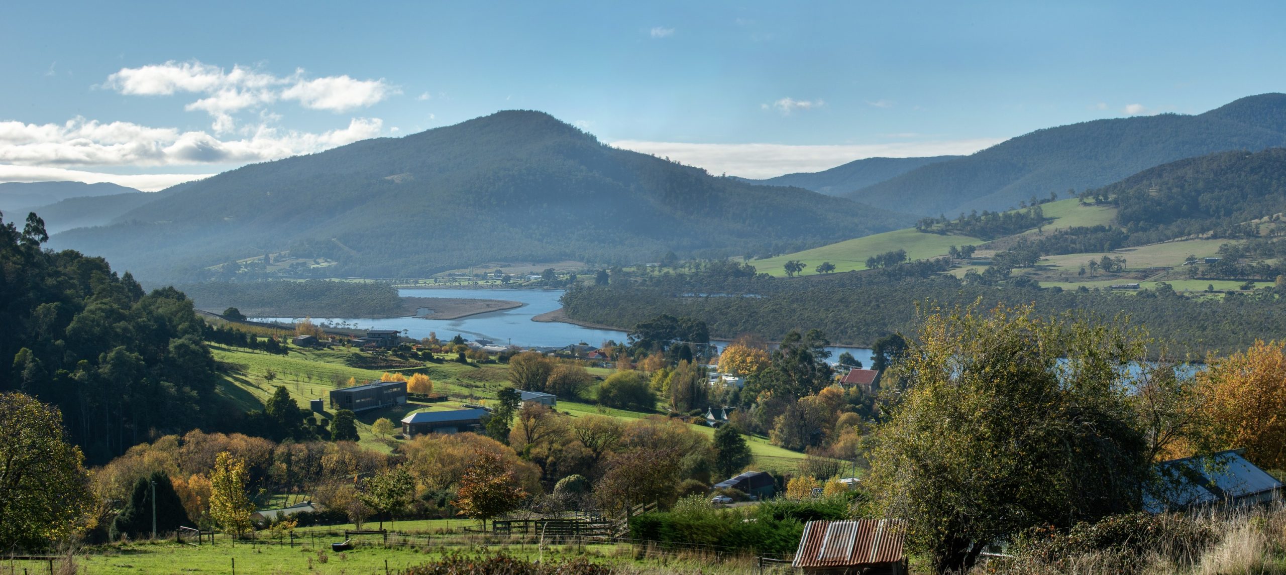 A scenic landscape of a lush valley with rolling hills, a river winding through the middle, and scattered houses. The trees are a mix of green and autumn hues, under a clear blue sky and soft clouds.