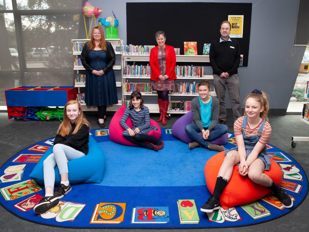 A group of people posing in a library. Four children sit on colorful beanbags arranged on an alphabet-themed rug. Three adults stand behind them, smiling. Shelves filled with books are in the background.
