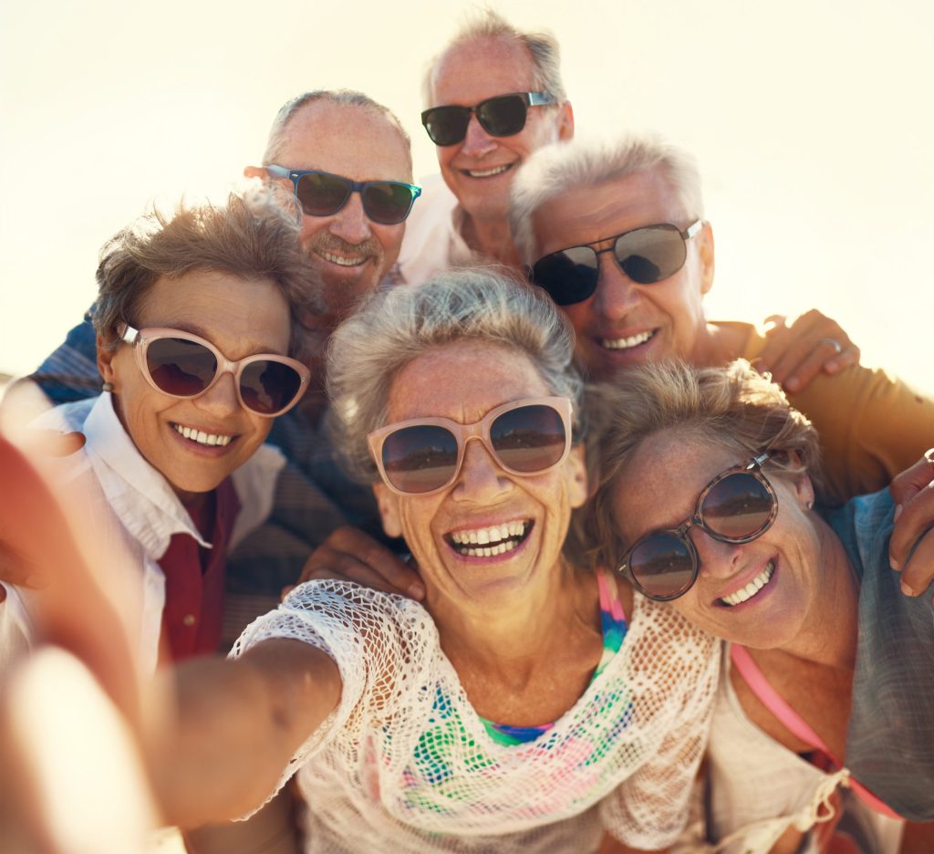 A group of six older adults is joyfully taking a selfie outdoors. They are all wearing sunglasses and smiling widely. The background is bright and sunny, suggesting a pleasant day.