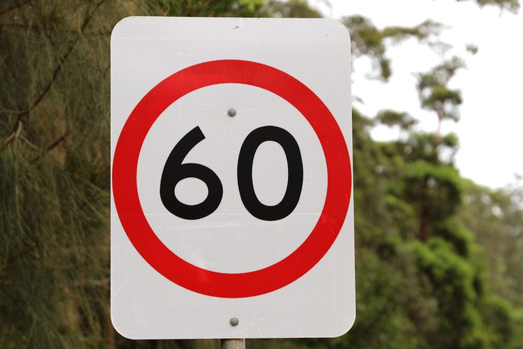 A speed limit sign with the number 60 inside a red circle, indicating a maximum speed of 60 kilometers per hour. Trees and greenery are visible in the blurred background.