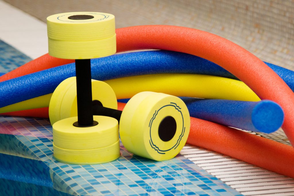 A pair of yellow foam dumbbells sits at the edge of a swimming pool, surrounded by colorful pool noodles in orange, red, yellow, and blue. The pool tiles are visible in shades of light and dark blue.