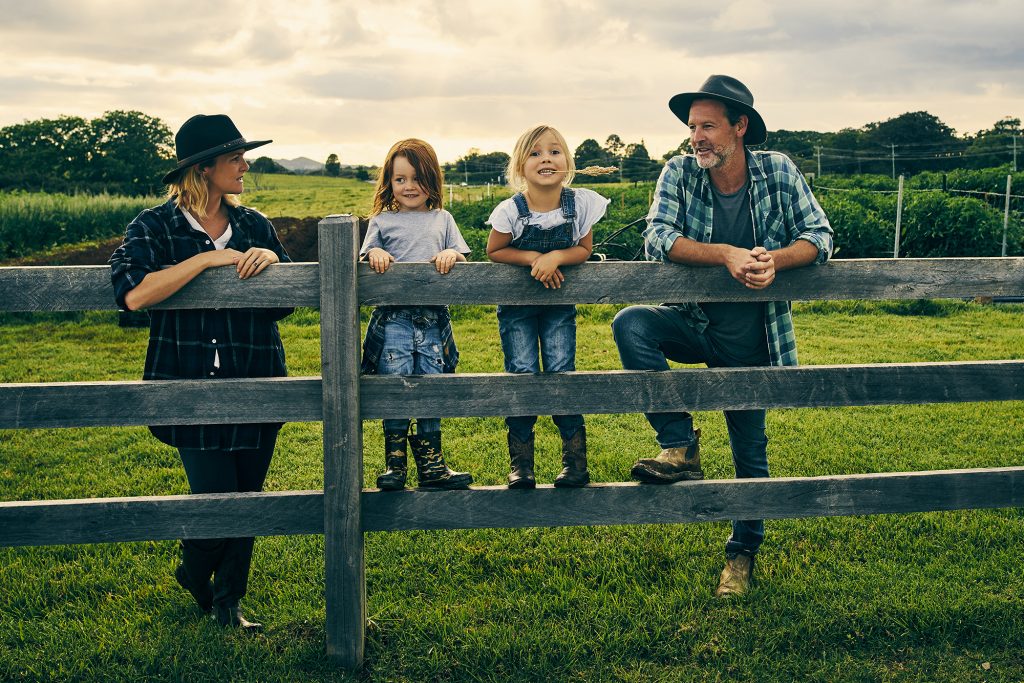 A family stands by a wooden fence in a grassy field. Two children are sitting on the fence, smiling. An adult is leaning on the fence, while another stands with arms crossed. The background shows green fields and a partly cloudy sky.