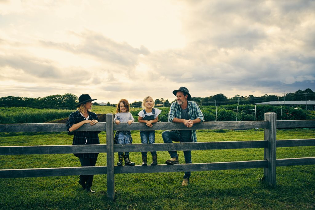 A family of four, two adults and two children, stand and sit on a wooden fence in a lush green field under a partly cloudy sky. They are smiling and wearing casual, countryside attire.