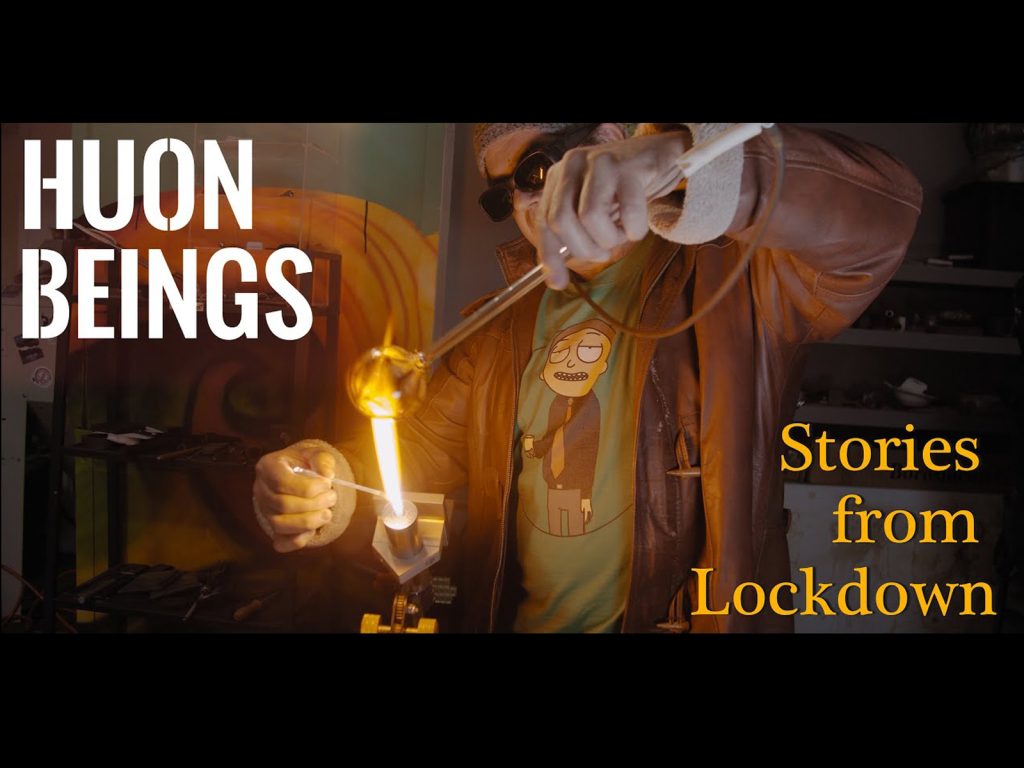 A person wearing sunglasses and a leather jacket is glassblowing, holding a glowing piece of glass with tools. Text on the image reads HUON BEINGS: Stories from Lockdown.