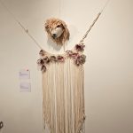 A wall-mounted textile art piece features a sheeps head at the top with long, hanging fringes of yarn below. The yarn is adorned with pink and purple flower-like decorations. The artwork is suspended from the ceiling by strings.