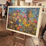 A vibrant painting of colorful flowers in a garden on display in an art gallery. The artwork is on an easel, surrounded by visitors observing and discussing the pieces. The gallery has wooden floors and exposed beams.
