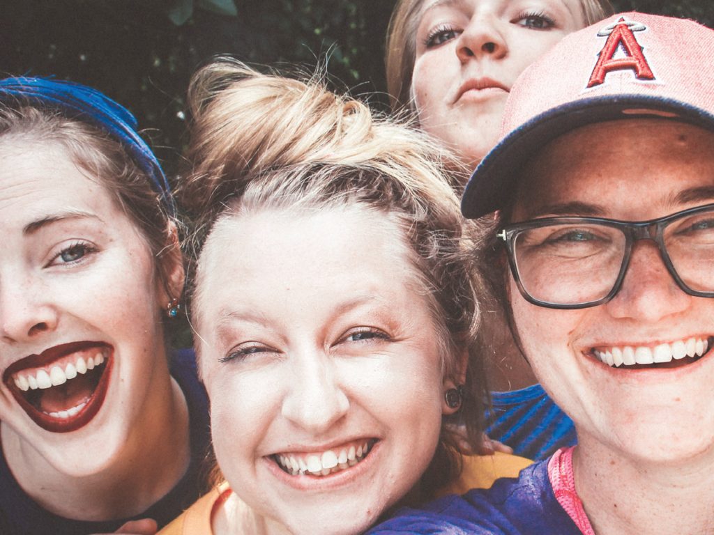 Four people smiling and taking a selfie outdoors. They are close together, showing expressions of joy. One is wearing glasses and a cap with an A logo. Another wears a blue headband. The group is lit by natural light.