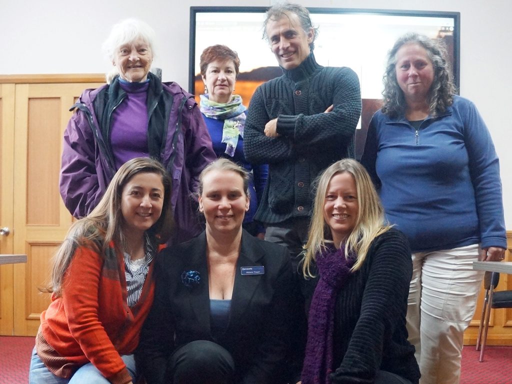 A group of seven people posing indoors, with a wooden door and a screen in the background. They are wearing casual and warm clothing, and all are smiling at the camera.