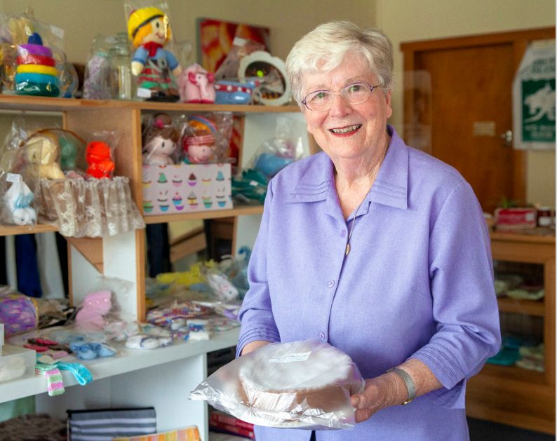 Elderly woman with short white hair smiling, holding a packaged cake, stands in a shop filled with colorful toys and handmade items on shelves. She is wearing a light purple blouse and glasses.
