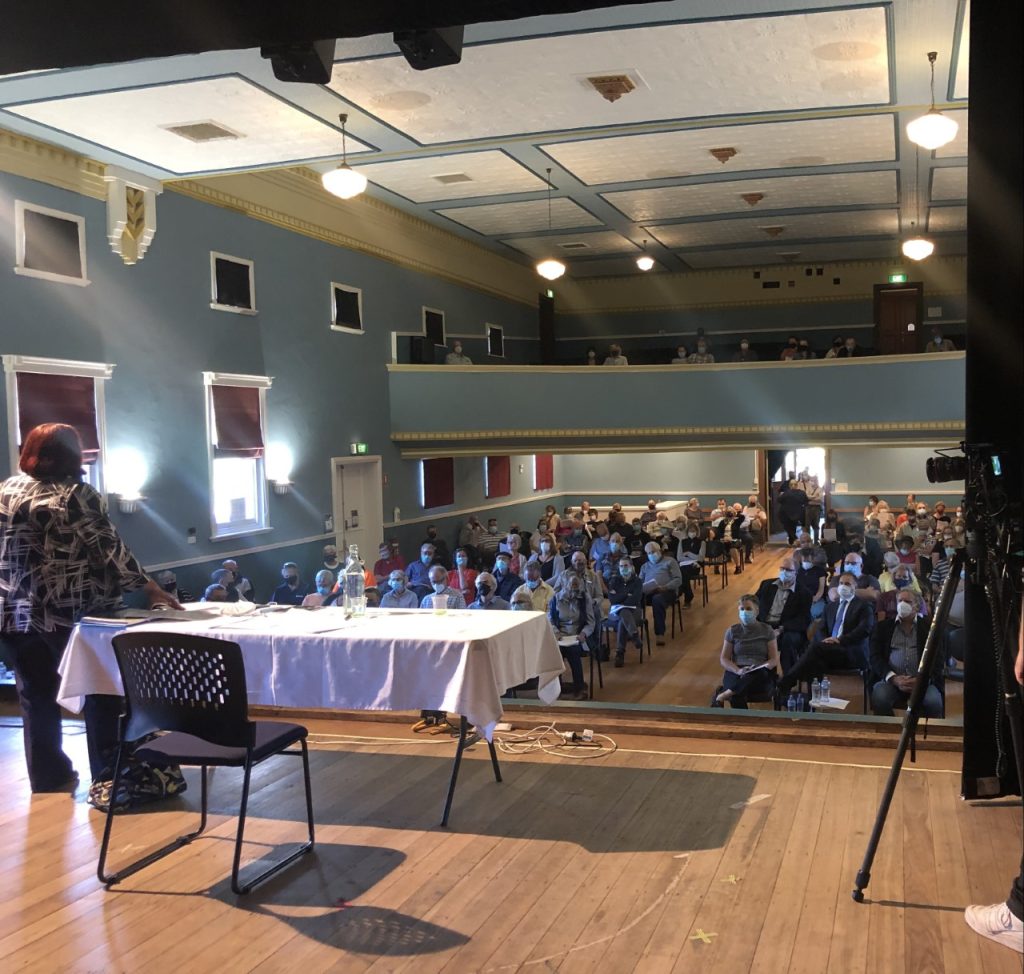 A person stands on a stage in front of a table with a microphone, addressing an audience seated in rows. The venue is a large hall with a light blue interior, featuring windows, balconies, and decorative molding. People fill the ground and balcony levels.