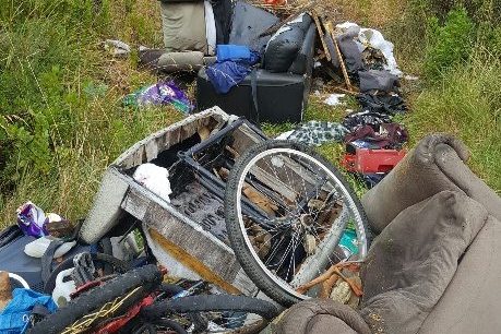 A pile of discarded trash in a grassy area, including broken furniture, a bicycle wheel, cushions, and various debris. The surrounding area is overgrown with grass and bushes.