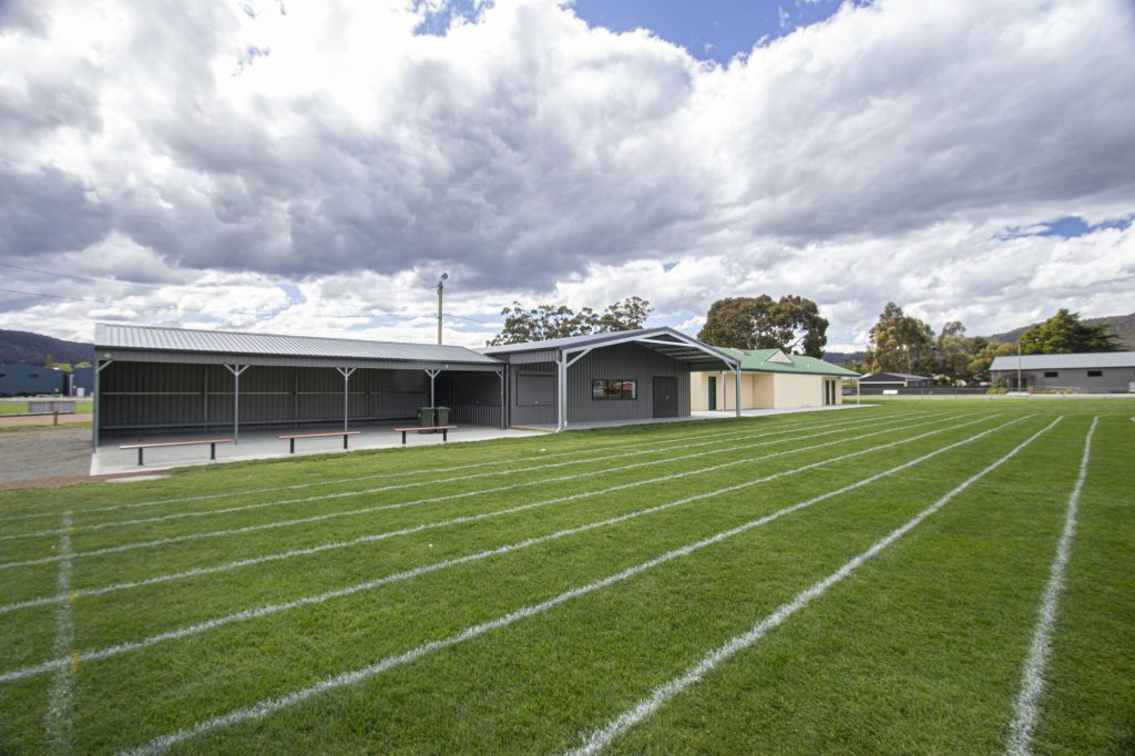 Sports field with a green grass running track, outlined by white lines, adjacent to a modern, grey pavilion. The sky is overcast with thick clouds, and trees are visible in the background.
