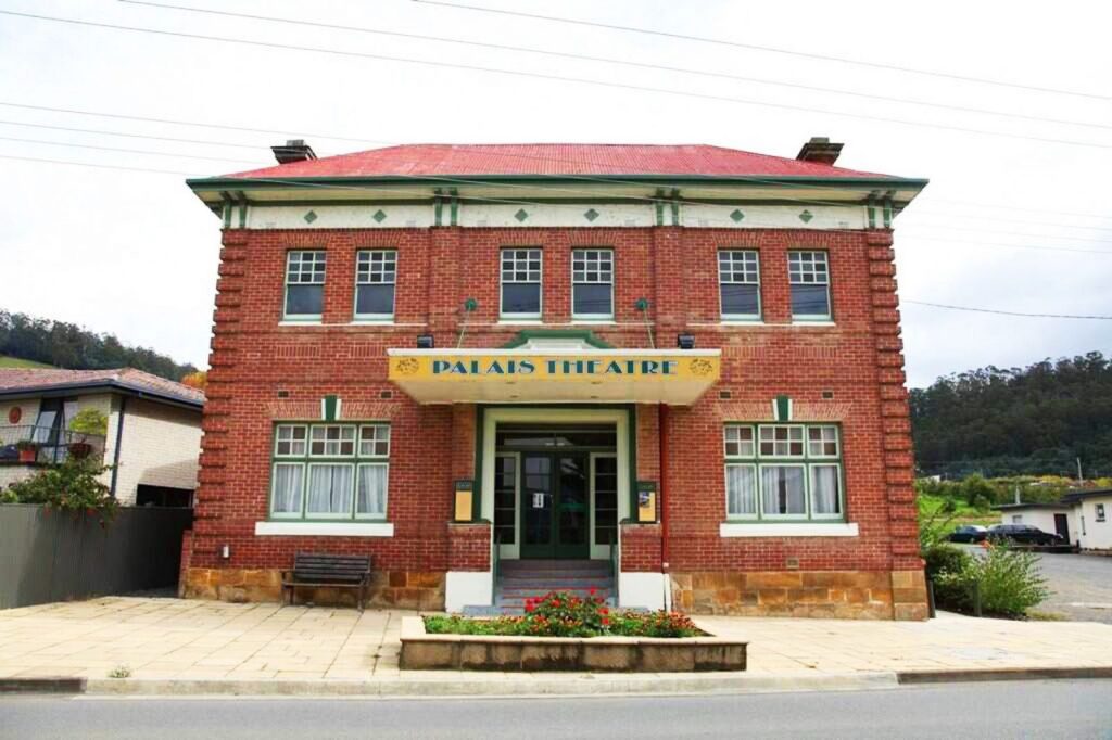A historic brick building labeled Palais Theatre with a red roof and symmetrical windows. The entrance has a small garden in front, and there’s a bench to the left. The setting is on a quiet street with a backdrop of trees.