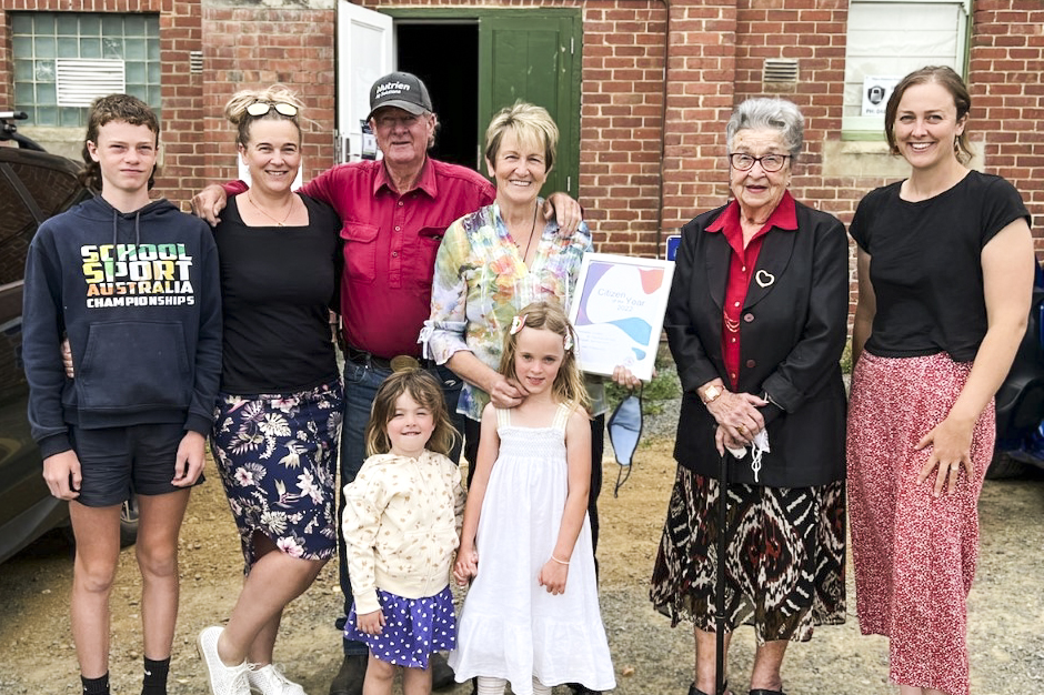A group of seven people, including adults and children, pose together outdoors in front of a brick building. They are smiling, with one holding a certificate. The group consists of three children and four adults.