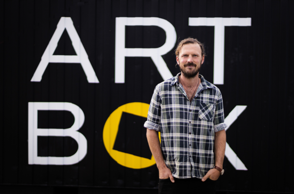 A person with a beard stands smiling in front of a black wall with ART BOX written in large white letters. They are wearing a plaid shirt, and their hands are in their pockets.