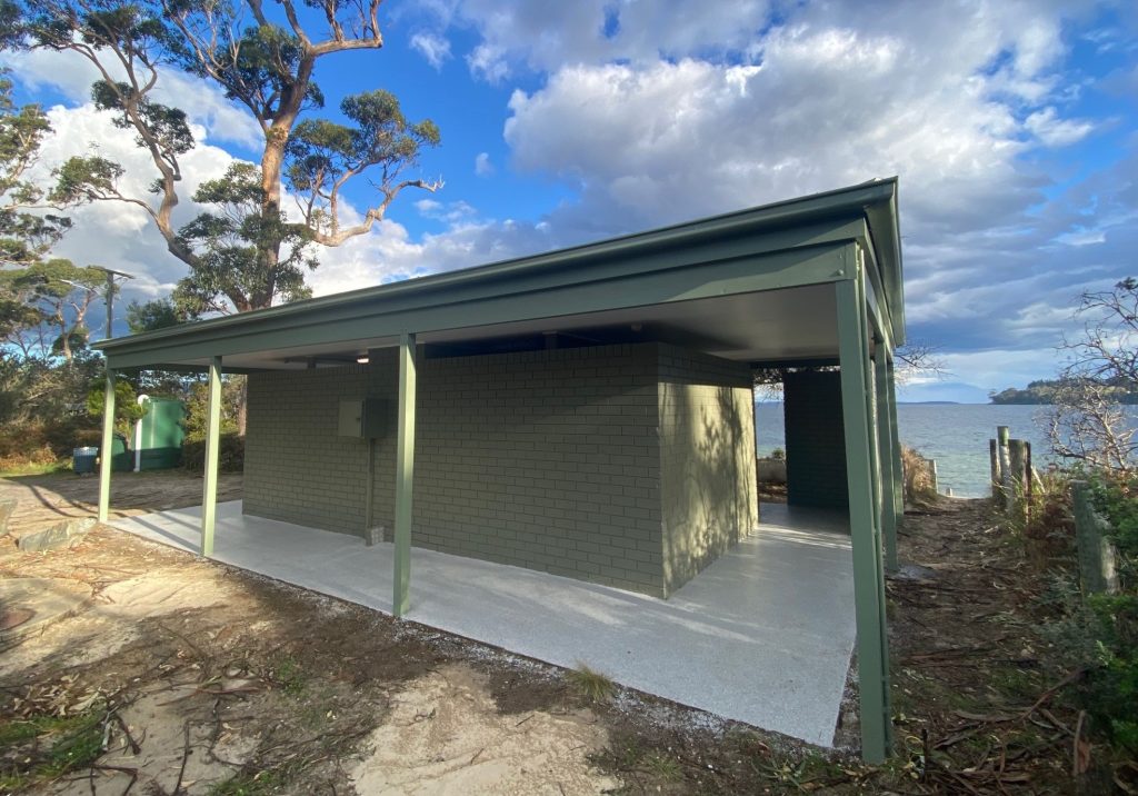 A green brick public restroom with a covered area, located near a body of water. Tall trees surround the building, and theres a partly cloudy sky above. The restroom is positioned on a sandy, leafy ground with a view of the water in the background.