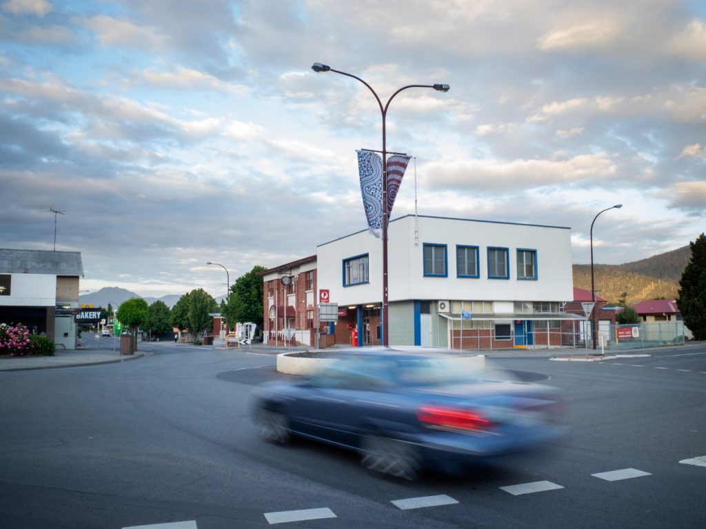A blue car speeds past a town intersection in the foreground. The background features a red and white building beside other structures. A cloudy sky and distant hills are visible. The scene is calm with little activity.
