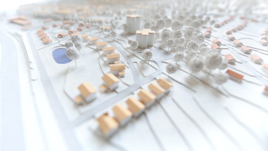 A detailed architectural model displays a small neighborhood with houses, trees, and a pond. Paths crisscross the area, with a focus on building placement and greenery. The model uses soft colors, giving a peaceful, structured appearance.