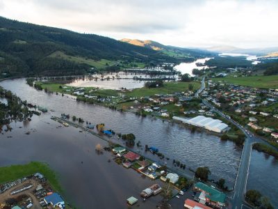 Aerial view of a flooded landscape with submerged houses and roads. Green hills in the background surround the inundated area, where water stretches over fields and residential areas. Partially submerged trees and structures are visible.
