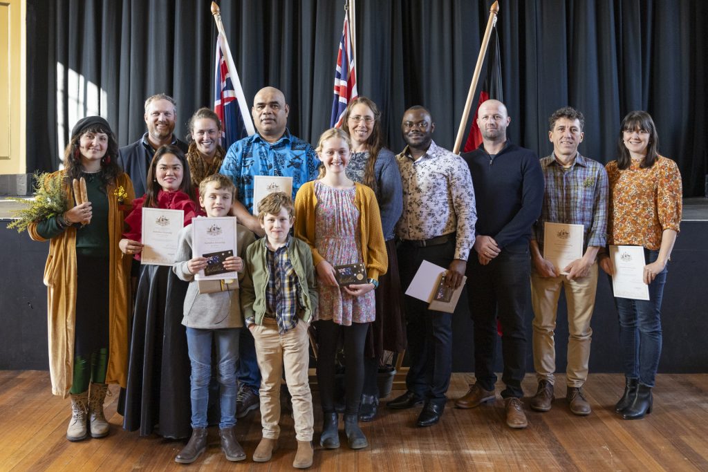 A group of diverse individuals, including men, women, and children, stand on a stage holding certificates. They are smiling, with two flags displayed in the background. The setting appears formal, likely a ceremony or event.