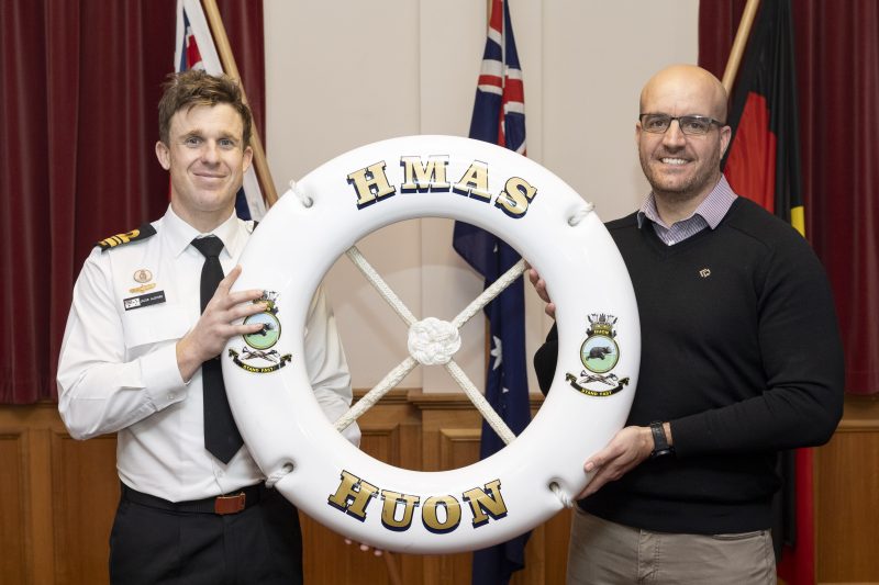 Two men are holding a lifebuoy with the text HMAS HUON. The man on the left is wearing a white naval uniform with badges. The man on the right is in a black sweater. They are standing indoors, with Australian flags in the background.