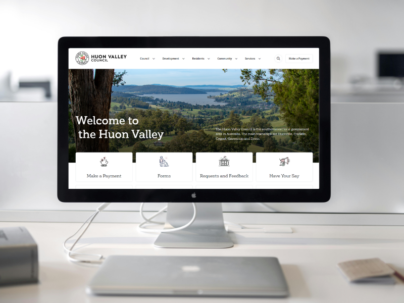 Computer screen displaying the Huon Valley Council website homepage, showcasing a scenic landscape with lush greenery and a river. The page includes options for making a payment, accessing forms, sending feedback, and staying informed.