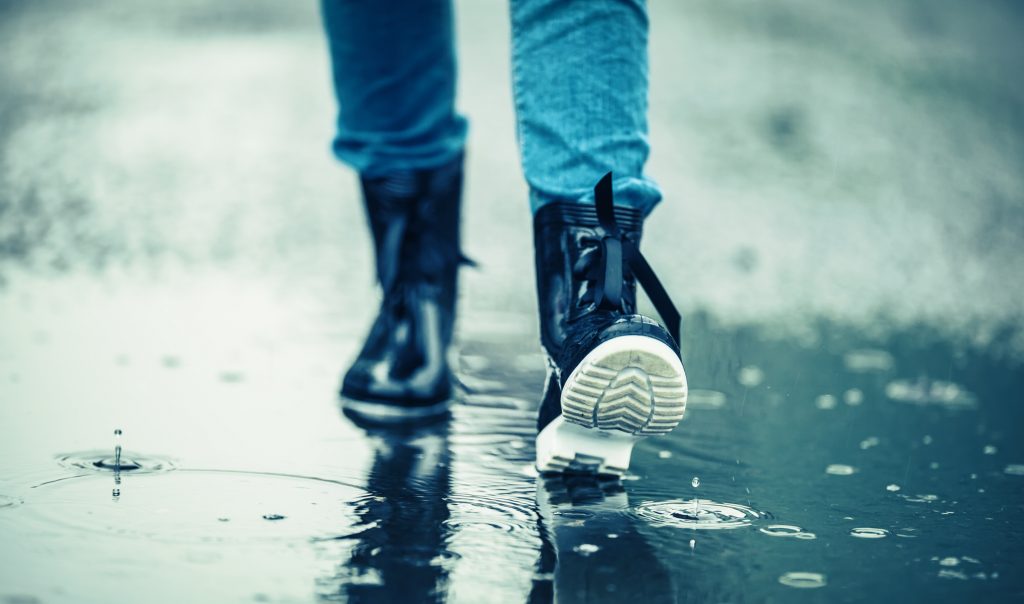 A person wearing shiny black boots walks through a puddle, creating small ripples in the water. The legs are dressed in blue jeans, and the scene is captured from the back, focusing on the person’s feet.