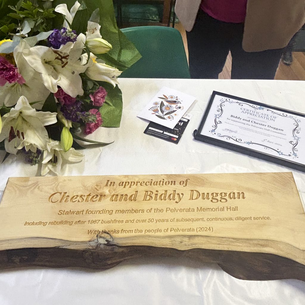 A wooden plaque with engraved text sits on a table. Beside it are a bouquet of flowers and a framed certificate. The plaque honors Chester and Biddy Duggan for their contributions to Pelverata Memorial Hall.
