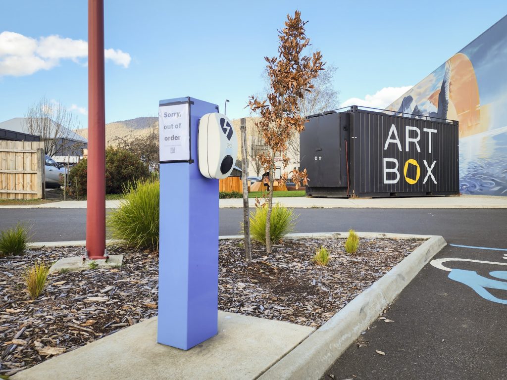 A charging station with a sign reading Sorry, out of order is situated in a parking area. A mural on a shipping container labeled ART BOX is seen in the background, surrounded by greenery and a clear sky.