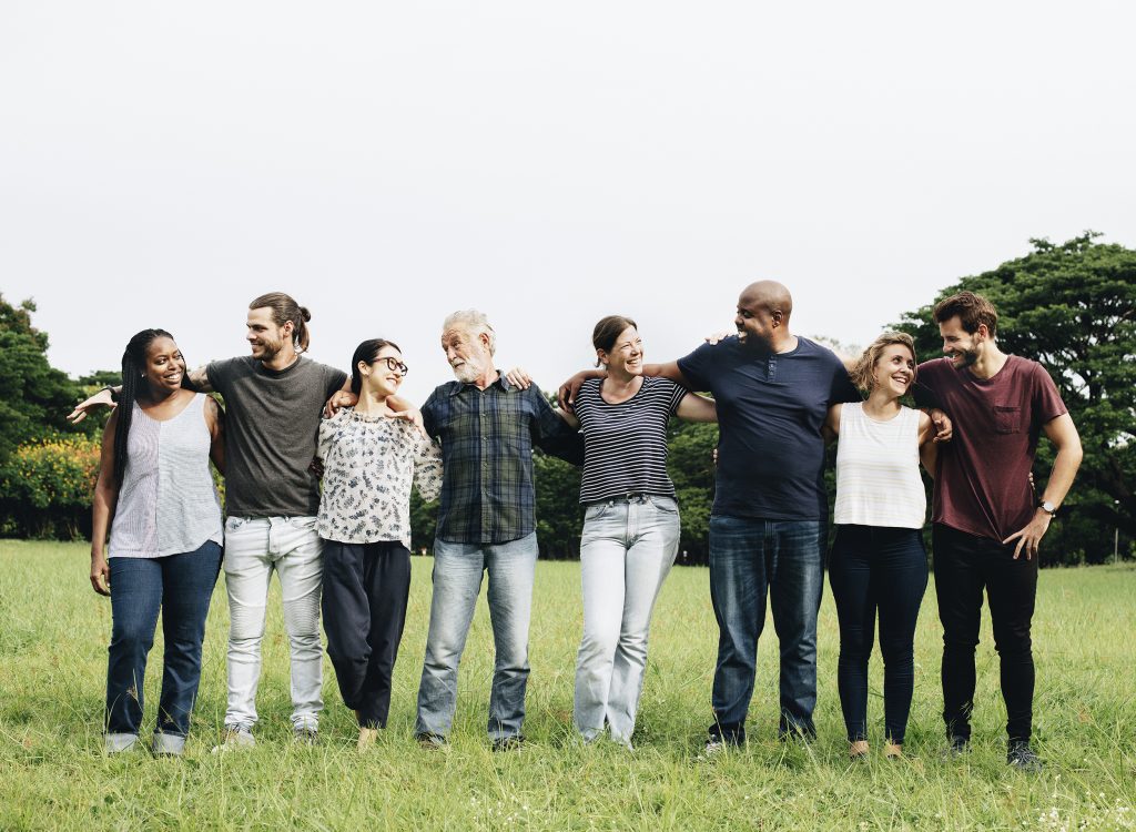 A diverse group of eight people stand in a row outdoors on grass, smiling and with arms around each others shoulders. Trees are visible in the background under a clear sky.