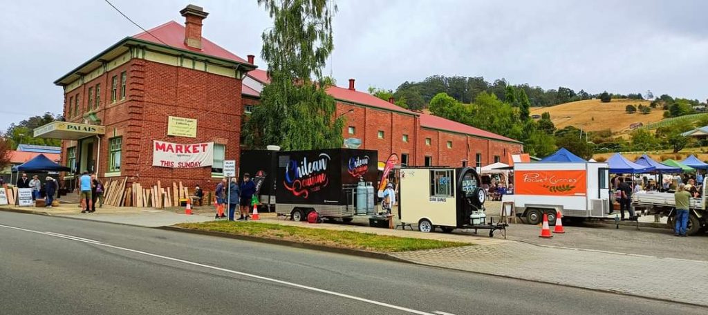 A street scene with a market outside a red brick building. Food trucks and stalls line the sidewalk with people walking around. A sign reads Market. Hills and trees are visible in the background under a cloudy sky.
