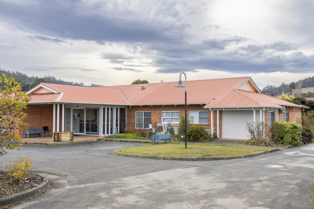 A single-story red brick building with a red roof, large glass entrance, and surrounding grassy area. A streetlamp stands in the foreground with a cloudy sky above. Bushes and a paved driveway are also visible.