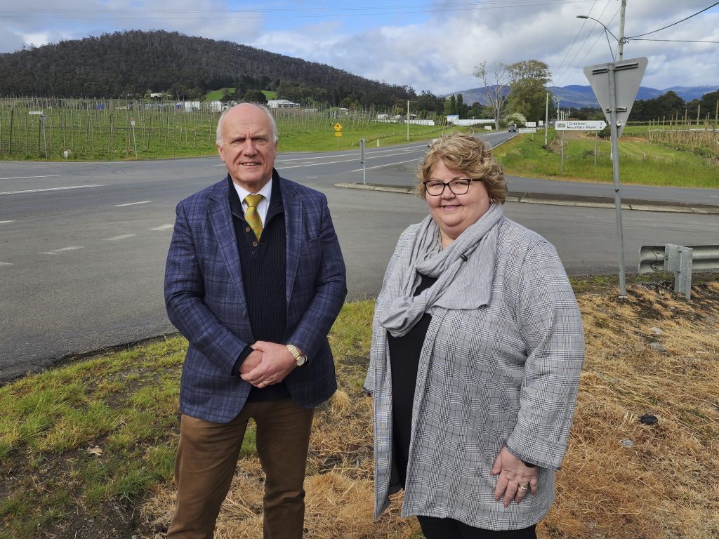 Two people stand on a roadside with hills and vineyards in the background. The man wears a blue plaid jacket, and the woman wears a gray plaid coat. Its a cloudy day, and the setting appears rural.