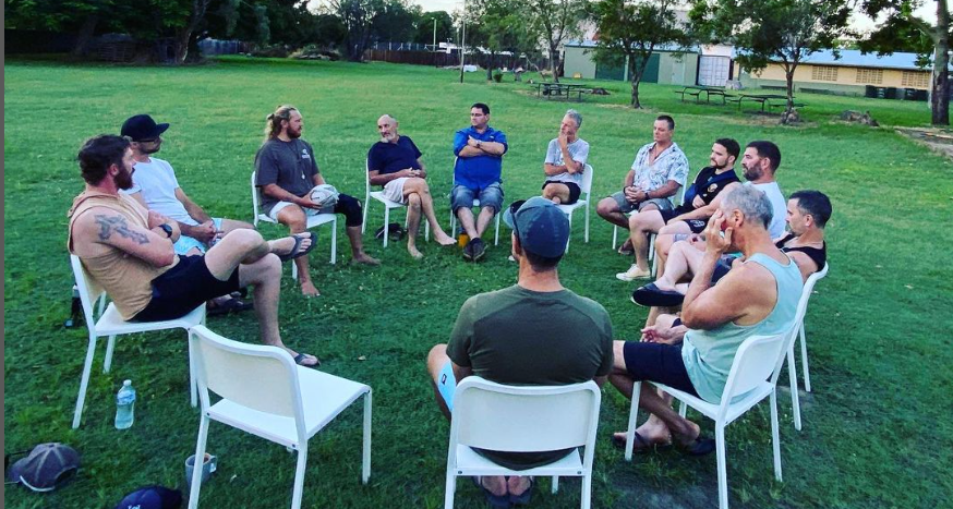 A group of men sitting in a circle on white chairs outdoors on a grassy field. They appear to be engaged in conversation, with trees and buildings visible in the background.