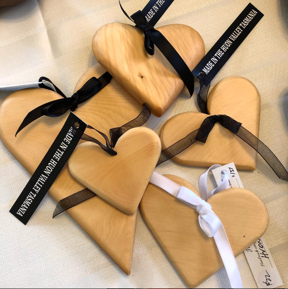 Six smooth, light wooden heart-shaped ornaments are arranged on a cream fabric, each tied with a bow made of black or white ribbon labeled “Made in the Huon Valley, Tasmania.” A handwritten tag is partially visible.