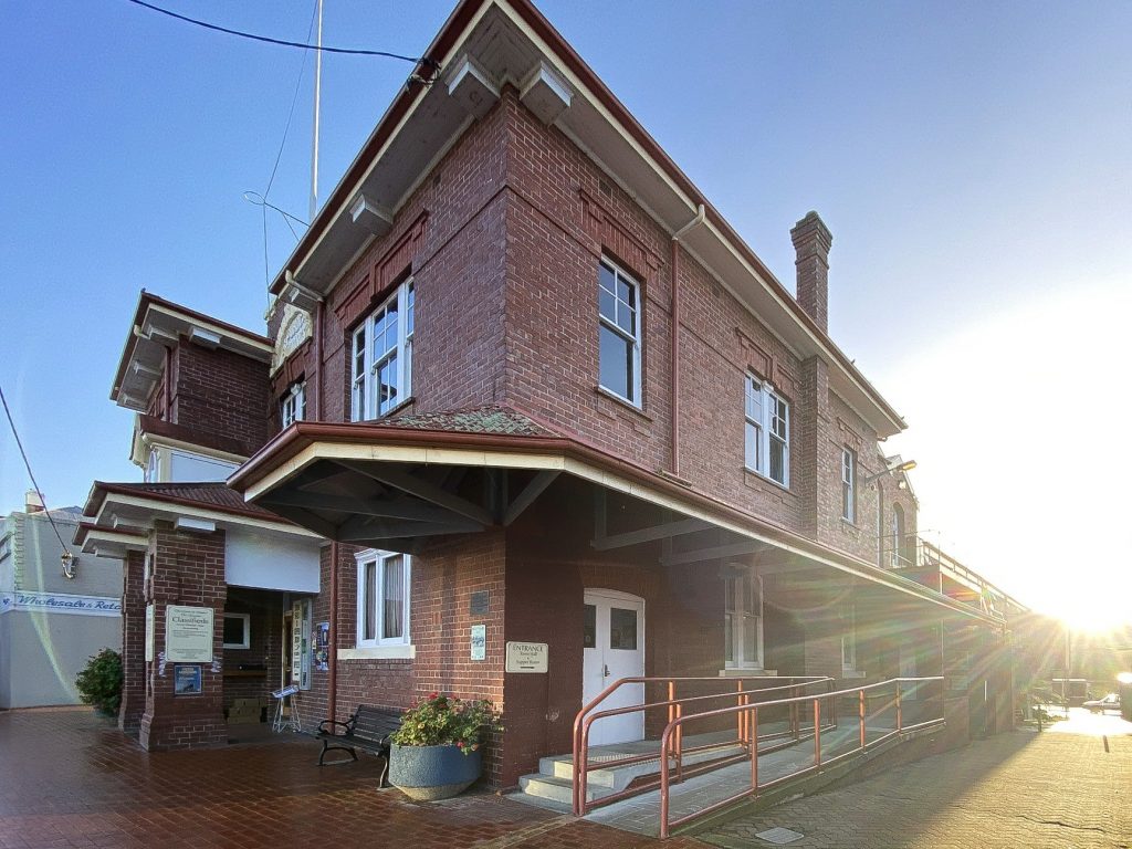 A historic brick building with a corner entrance. It features large windows, a sloped awning, and a ramp with a handrail. The scene is lit by early morning or late afternoon sun, casting long shadows on the brick-paved sidewalk.