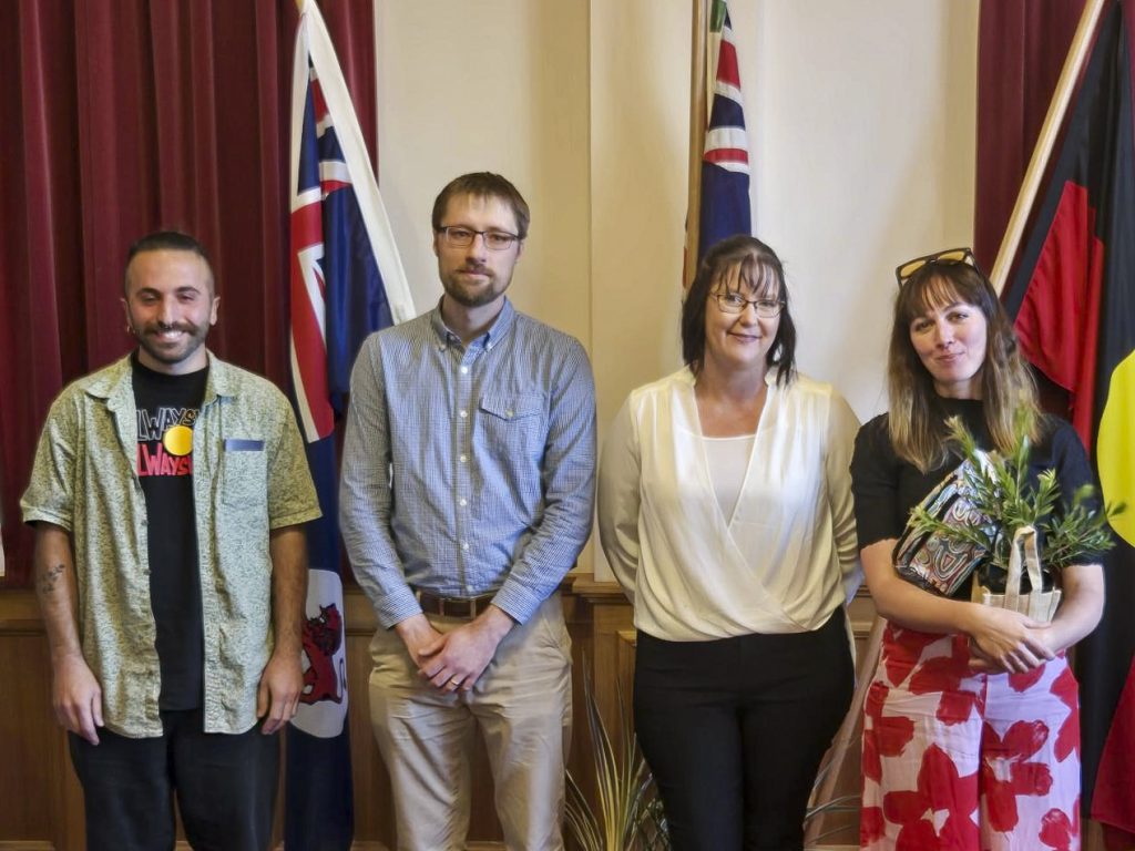 Four people stand in front of three flags. They are smiling and casually dressed. One person holds a potted plant. The flags include Australian and Aboriginal designs. The group is standing indoors on a wooden floor with red curtains behind them.