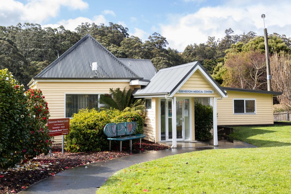 A small, yellow medical center with a metal roof is surrounded by greenery. A sign reads Geeveston Medical Centre. Theres a bench outside, and trees fill the background under a cloudy sky. A paved path leads to the entrance.